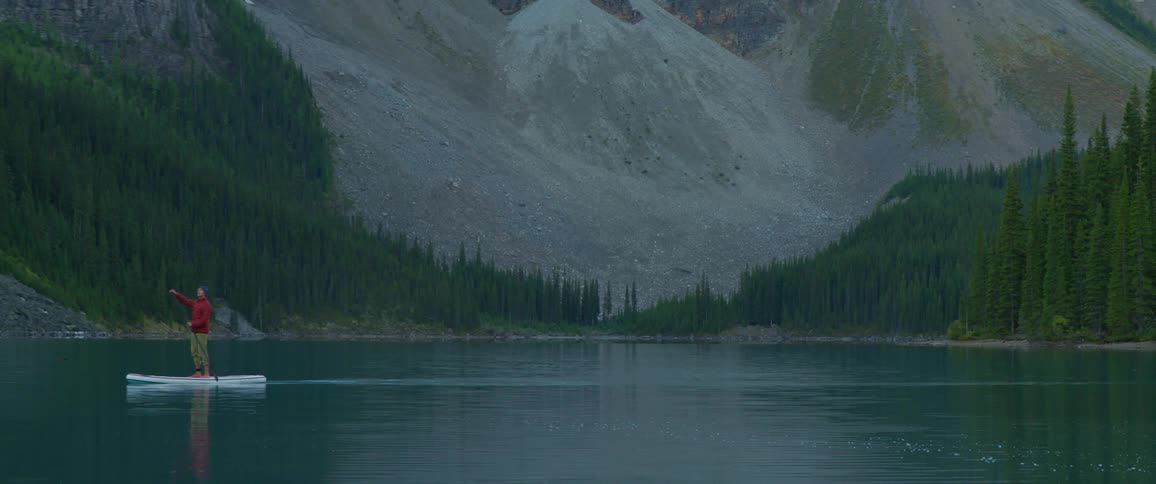 A person standing on a paddle board paddles across a lake at the base of a mountain