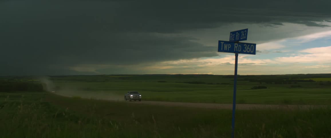 A truck travels down a dirt road through the countryside under a sky filled with dark clouds