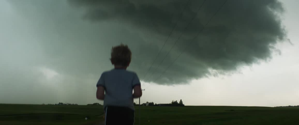A young boy walks alone in a rural field area with dark storm clouds above