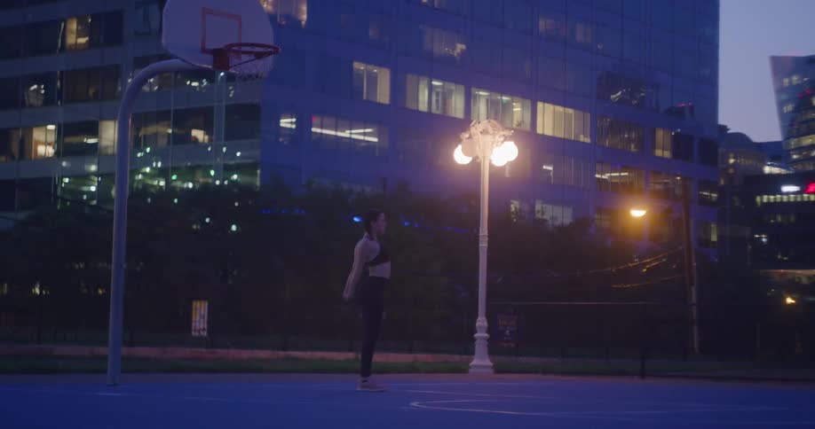 A woman in fitness clothing stretches out in a basketball court in a city at night