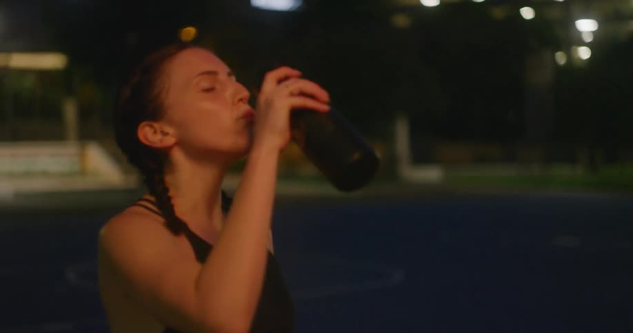 Woman rests after exercising and takes drink of water near city skyscrapers at night