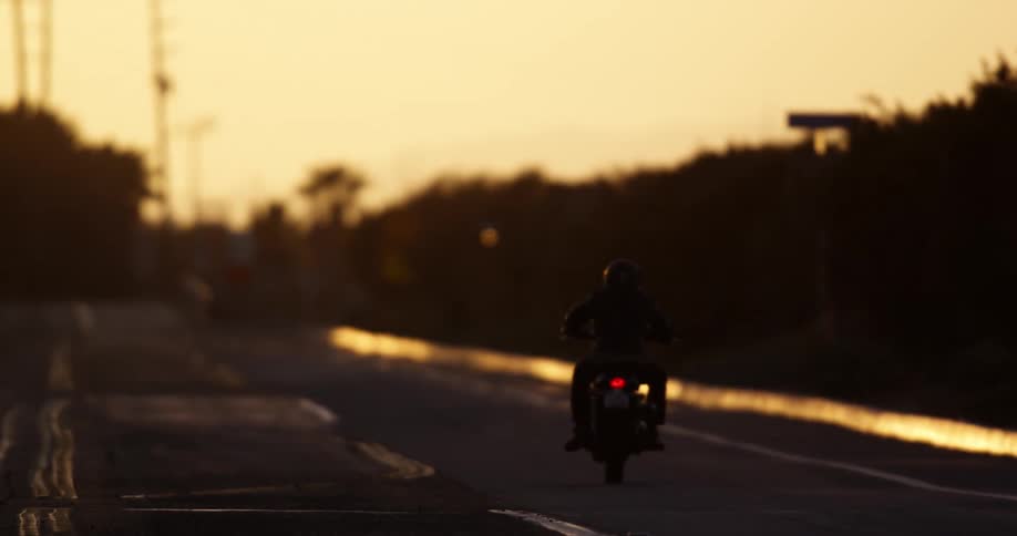 The silhouette of a motorcyclist can be seen as they are driving down a road at sunset