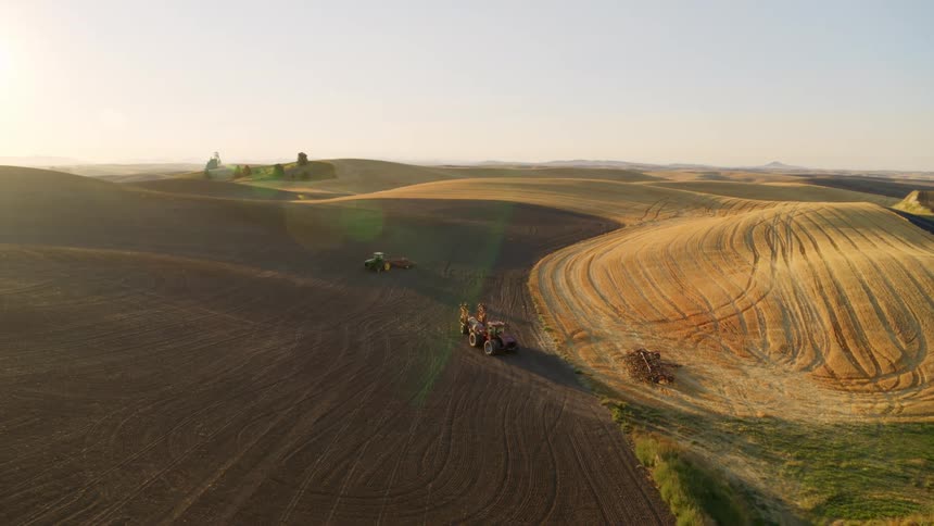 Flying over parked tractors and idle farm equipment on a freshly plowed field at sunrise