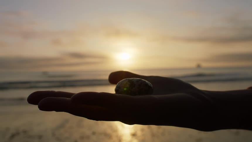 A person on the beach holding a stone in their hand with the sky and clouds in the background