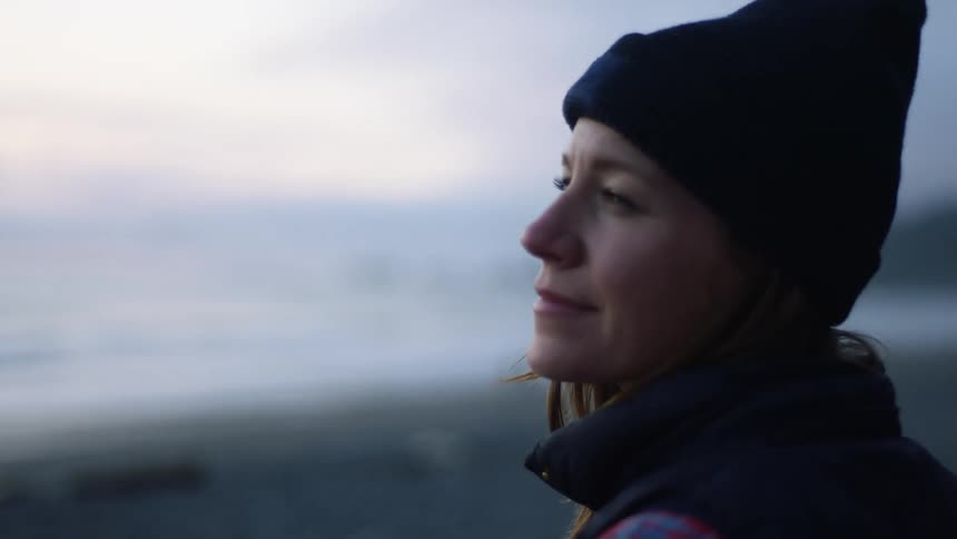 A woman standing on the beach smiles as she looks down the shore