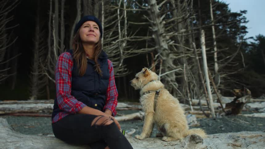 Woman with brown hair a black beanie and ripped pants sits on a tree trunk next to her dog in the woods
