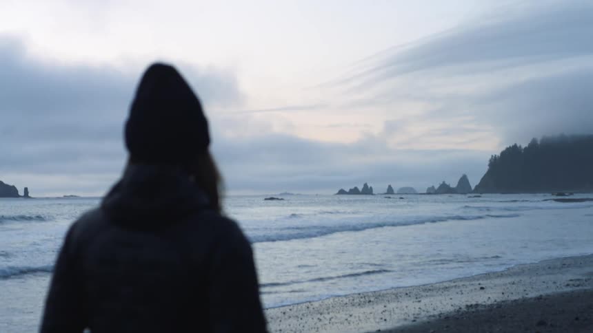 A woman wearing warm clothing walks along a coastal shoreline on a cold day