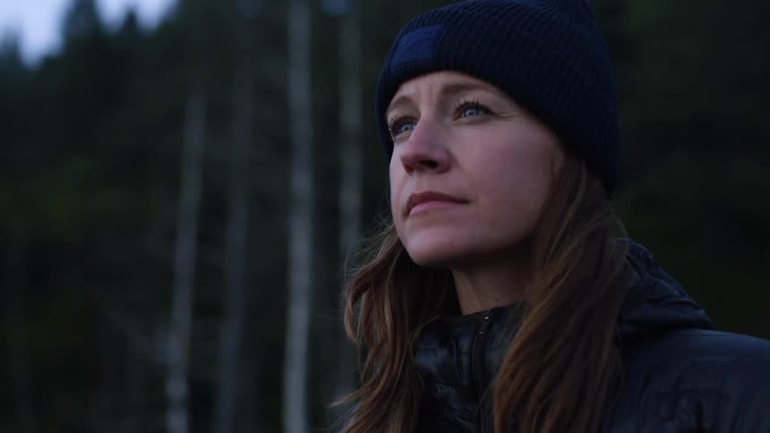 A middle aged woman dressed for cold weather stands outdoors near a woodland in the evening