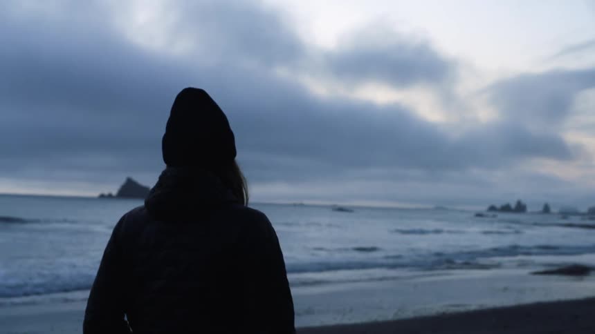 A woman in cold weather clothing walks a pet along the beach on a cloudy evening