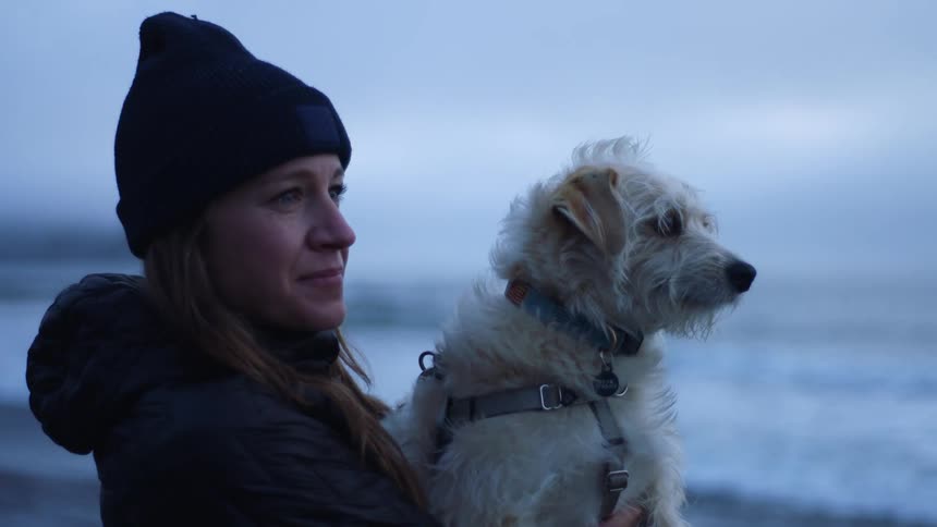 A woman holds her small white dog as they stand on the beach by the ocean