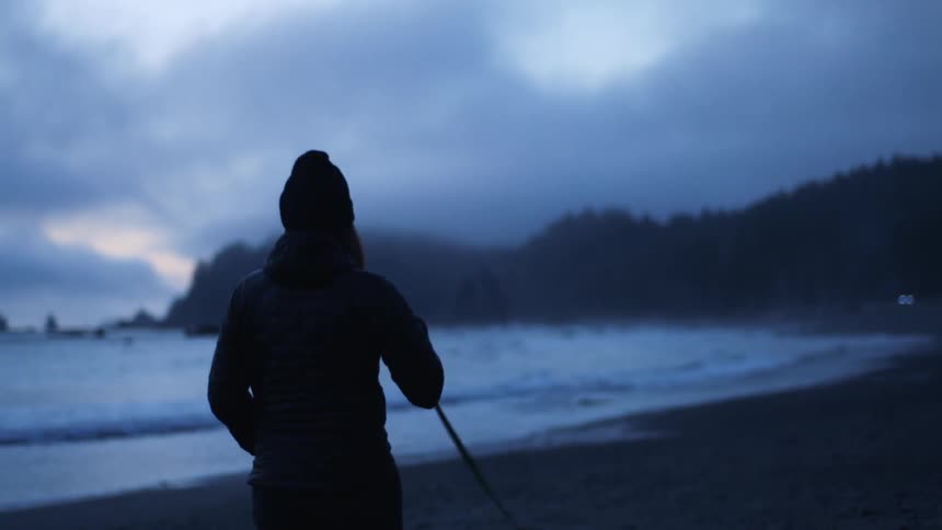 Woman wearing cold weather gear walks dog along beach