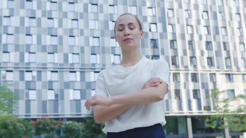 Woman with a white blouse and black pants crosses her arms and faces the camera with eye contact in front of office buildings