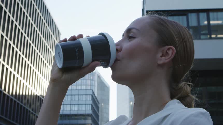 A woman stands outside an office building in a city and drinks a coffee