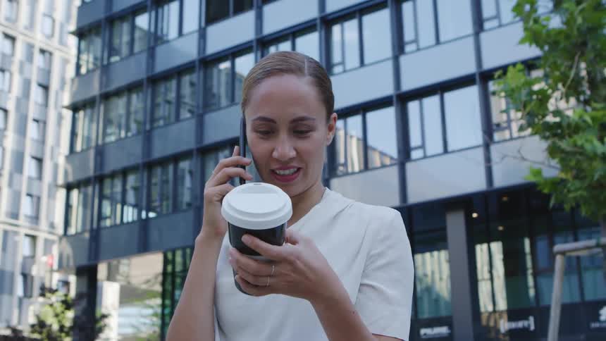Woman with a white blouse holds a plastic coffee cup and talks on a smartphone in front of the office buildings