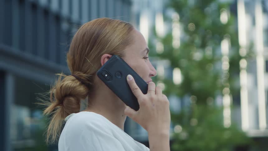 A woman smiles and laughs as she stands in front of a tall building talking on her smartphone