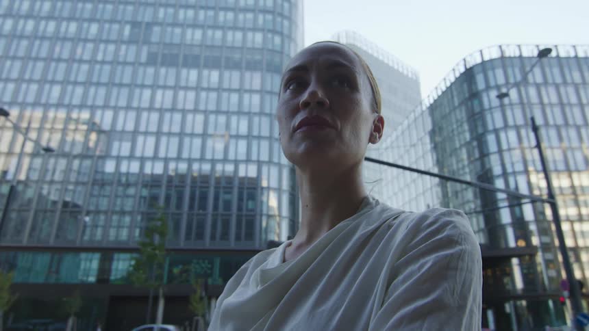 A blonde woman with her hair tied back is standing in the middle of city buildings as traffic drives past in the street behind her