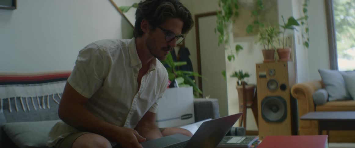 A man with glasses sitting on a couch next to a table and opening a laptop