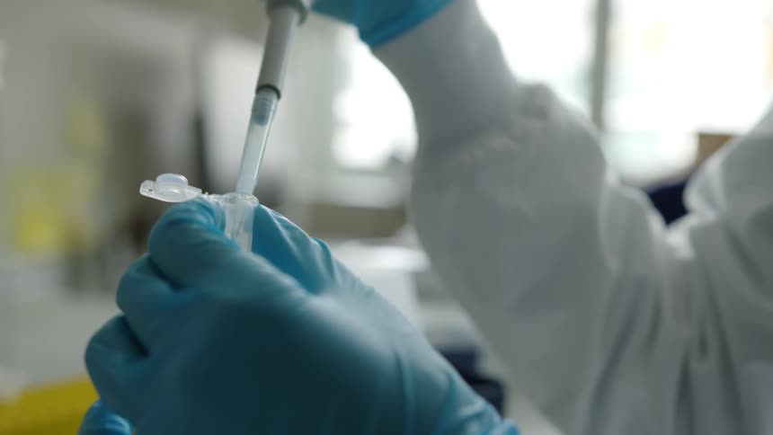 A person with a lab coat and gloves in a lab putting a liquid in a tube
