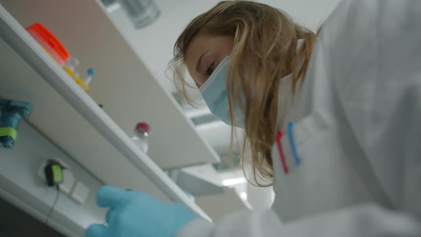A woman with blond hair wearing a face mask gloves and a white coat is injecting samples in a lab setting