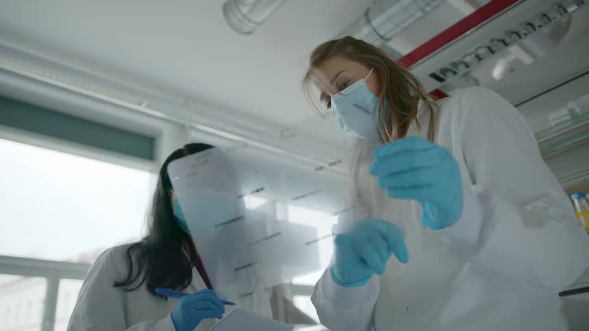 Two laboratory workers wearing masks and gloves examine a chart together
