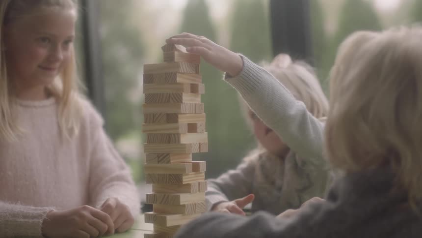 Three girls are sitting at a table playing a game inside with one girl placing a piece of wood with her hand
