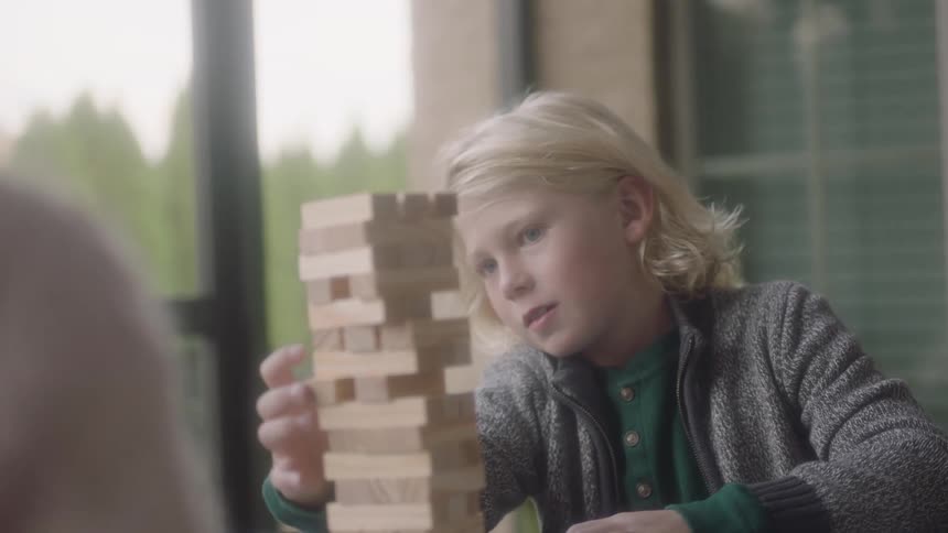 A blonde child is adjusting a piece of a wooden block in a game while sitting at a table on a balcony in the daylight