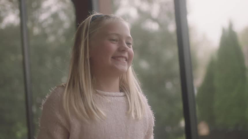 A blonde girl is expressive and smiling inside with windows looking out at trees during the day