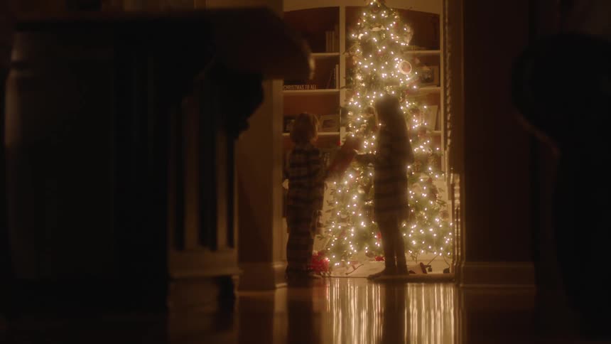 Children stand in front of a bright lit christmas tree shaking their presents and listening for whats inside