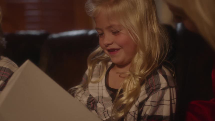 A young blonde girl smiles as she holds a present while sitting next to her mother on the couch