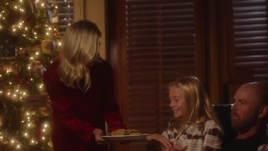 Dad sits with daughters on a couch in front of a christmas tree while a mom walks over to them and presents a plate of cookies