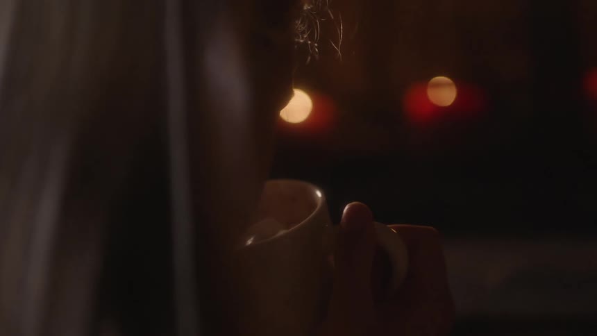 A person is sipping liquid out of a white mug at night with red candles in the room