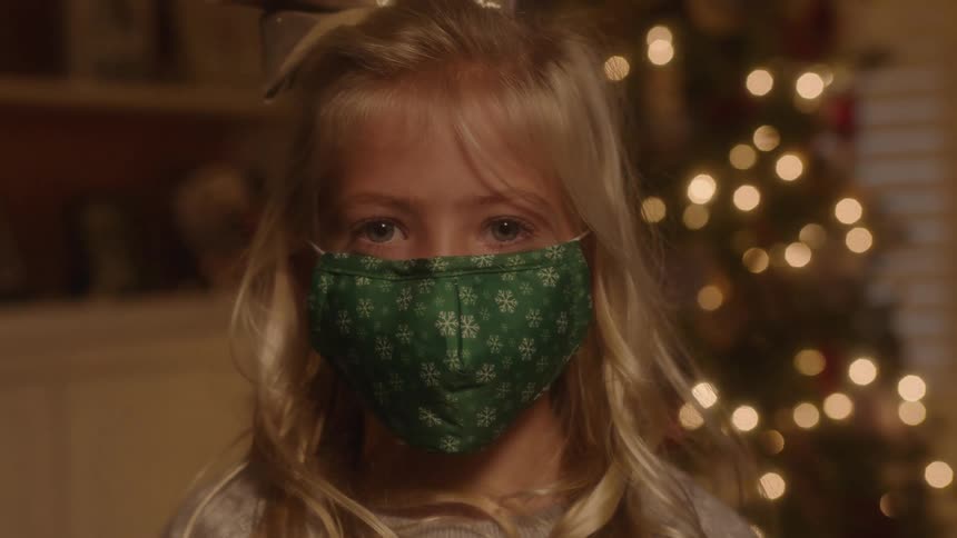 A girl with a mask in a room with a shelf and a christmas tree in the background