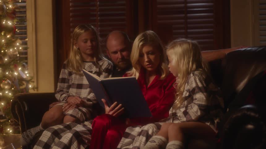 A woman sitting on a sofa with her husband and daughters reads a book aloud