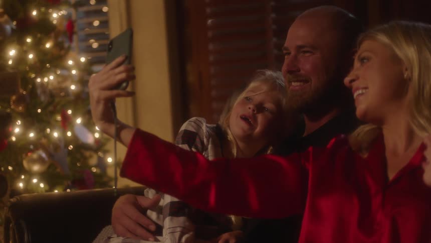 A man and woman huddle closely with their daughters taking a selfie by a christmas tree