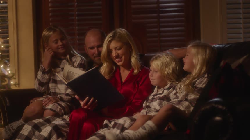 A family sits on the couch while the mom reads to the kids