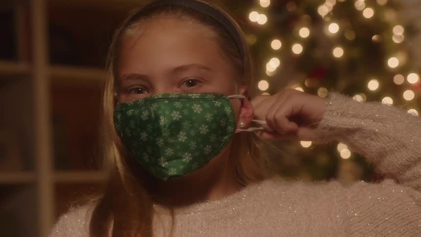 A blonde girl is smiling and placing a mask on her face while standing in front of christmas tree at night indoors