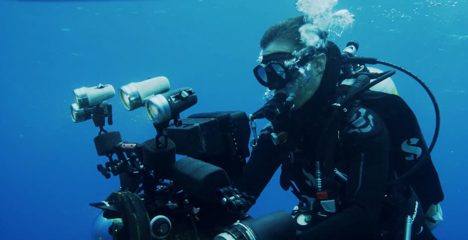 A scuba diver is underwater holding a camera with sunlight streaming from the waters surface