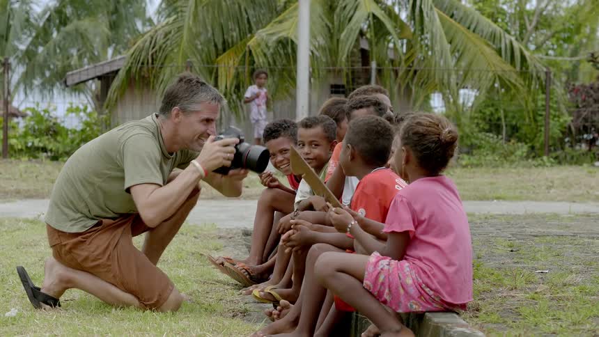 A man kneels down and takes photos of young children sitting on a rural or third world curb