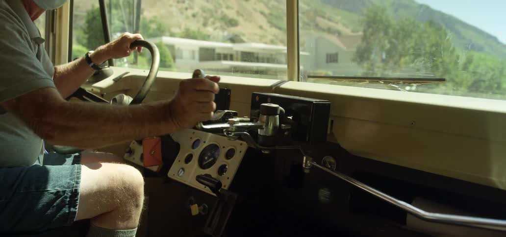 A man with a hat and a mask sitting in a vehicle pushing a lever with trees the sky and clouds in the background