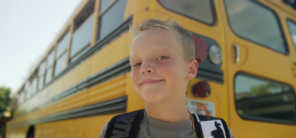 A blonde boy wearing a backpack stands next to a school bus outside in sunshine