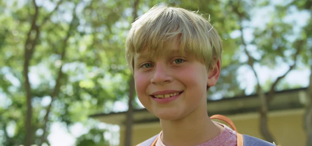 A young boy stands outside with a backpack on and smiles