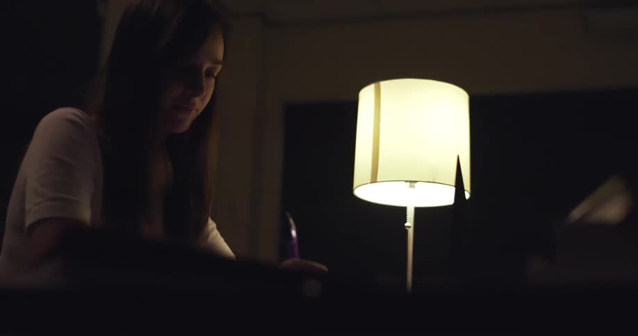 Girl with a white shirt and dark straight hair sits at desk and writes in a notebook next to a reading lamp in a dark room