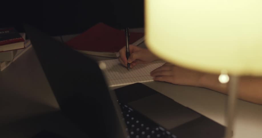 A woman sits at a desk with a laptop and writes on a sheet of paper