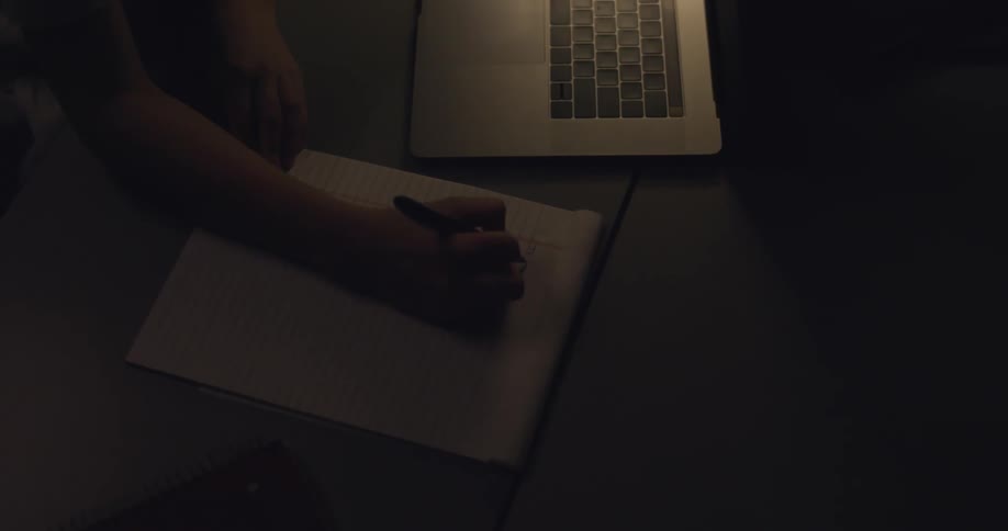 Girl writes in a notebook next to a laptop in the dark room