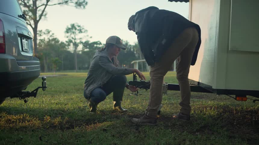 Couple works together to attach a trailer to a parked car in an open field