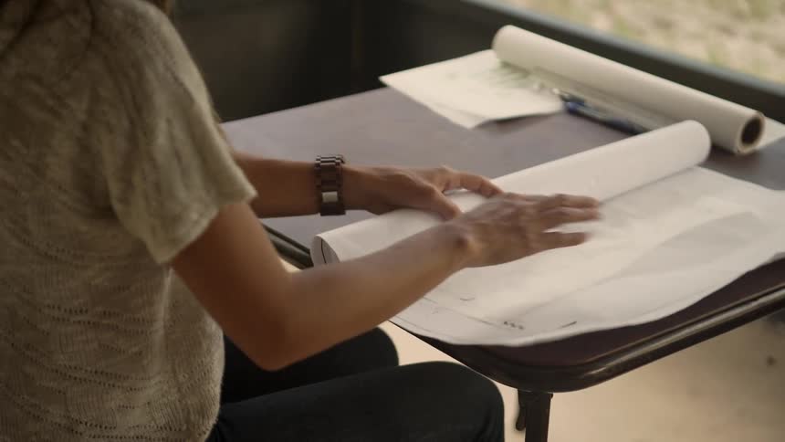 A woman with a wristwatch sitting at a table and opening a rolled up paper