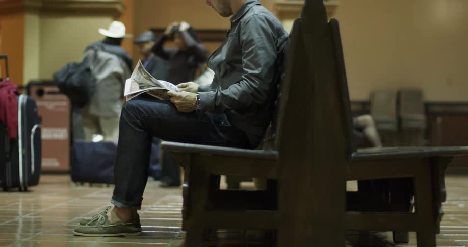 People pass time waiting at a train depot by reading newspapers and talking to other passengers