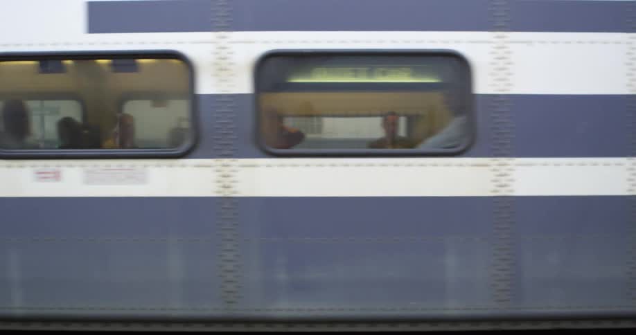 A purple and white train moves down the track with passengers visible through the windows