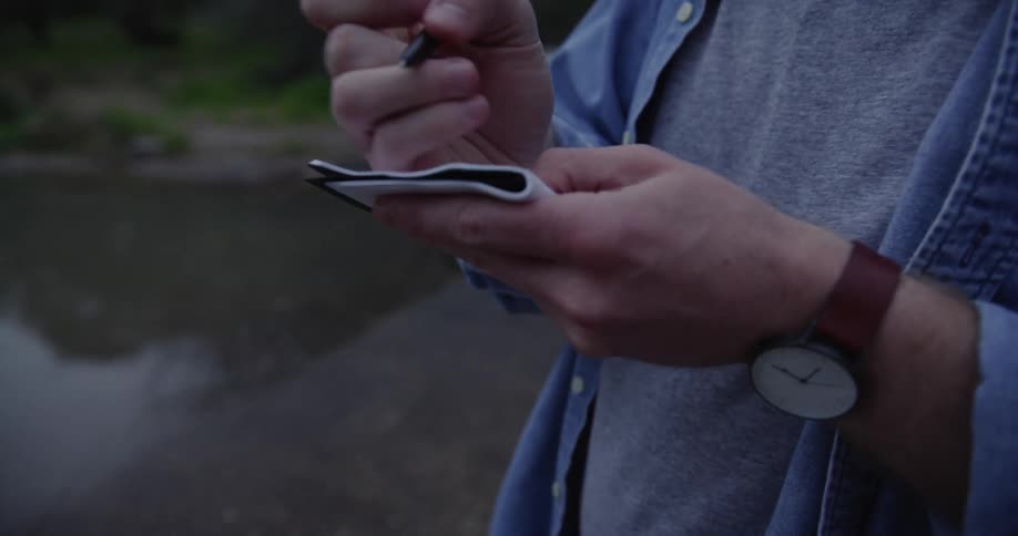 A man with a wristwatch writing on a pad with trees the sky and clouds in the background