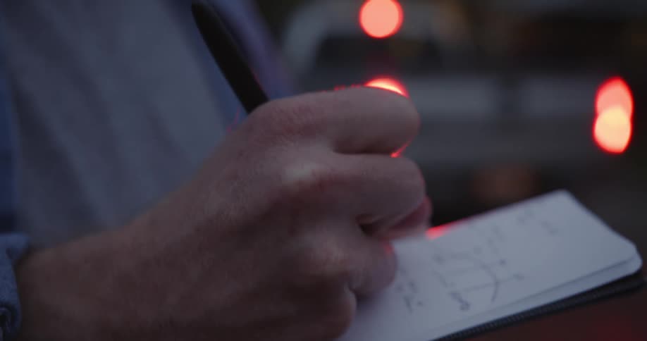 A man writing on a pad with a pen with a vehicle in the background